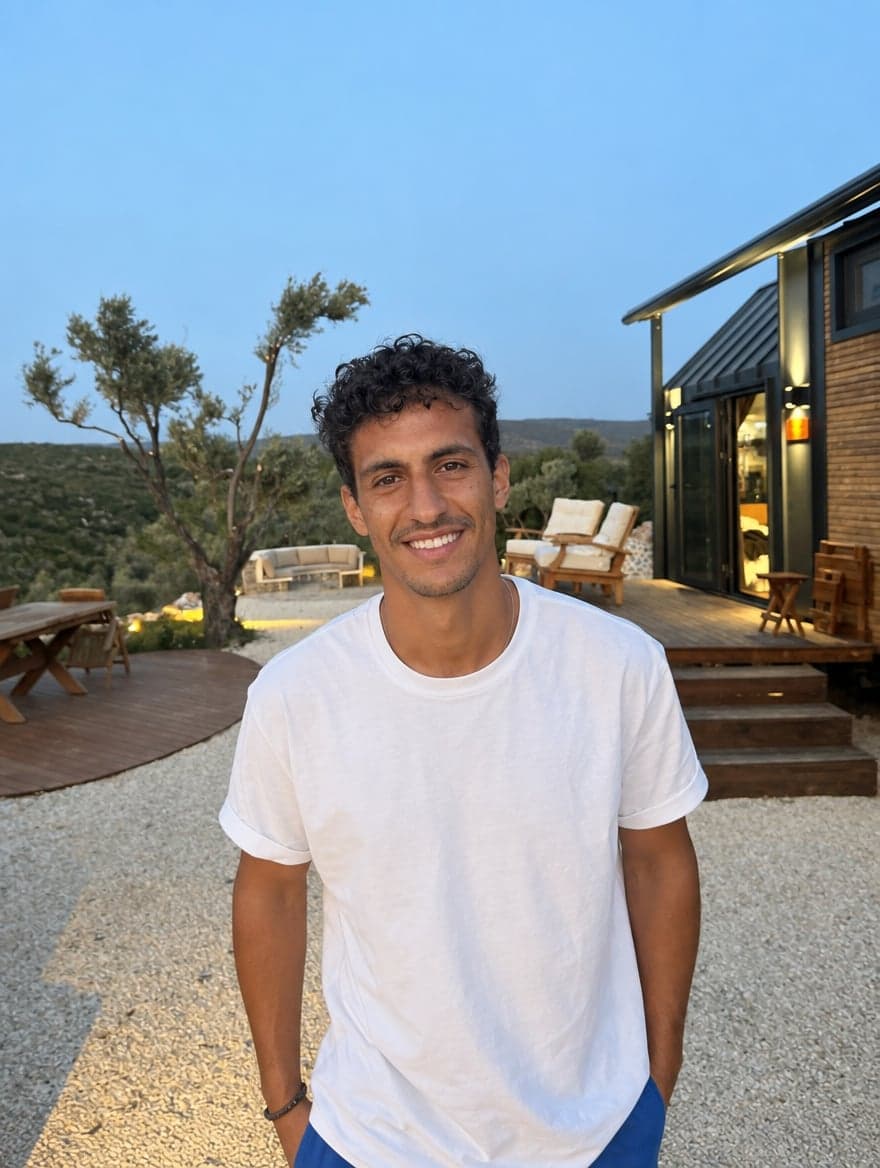 Man in a white T-shirt smiling outside a cabin at dusk