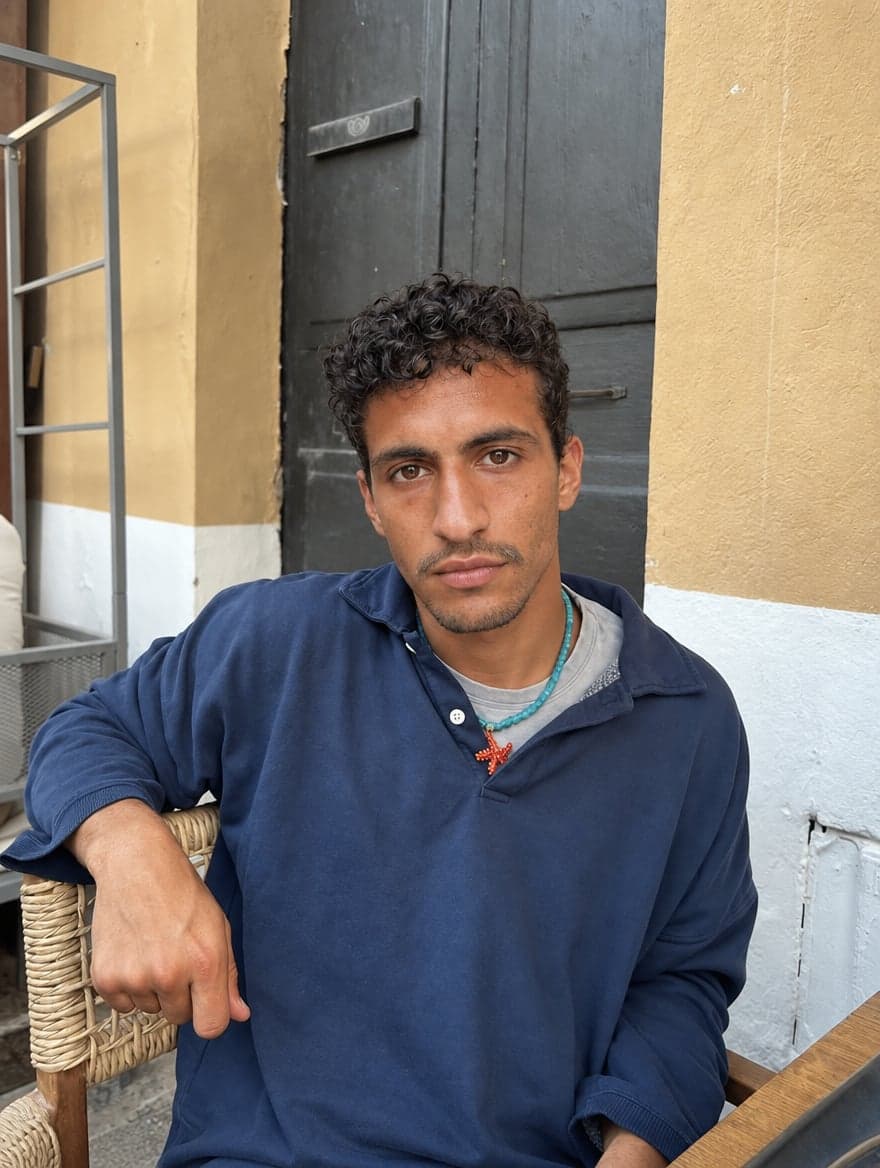 Man in a navy polo sweatshirt and beaded necklace sitting in a wicker chair by a yellow wall