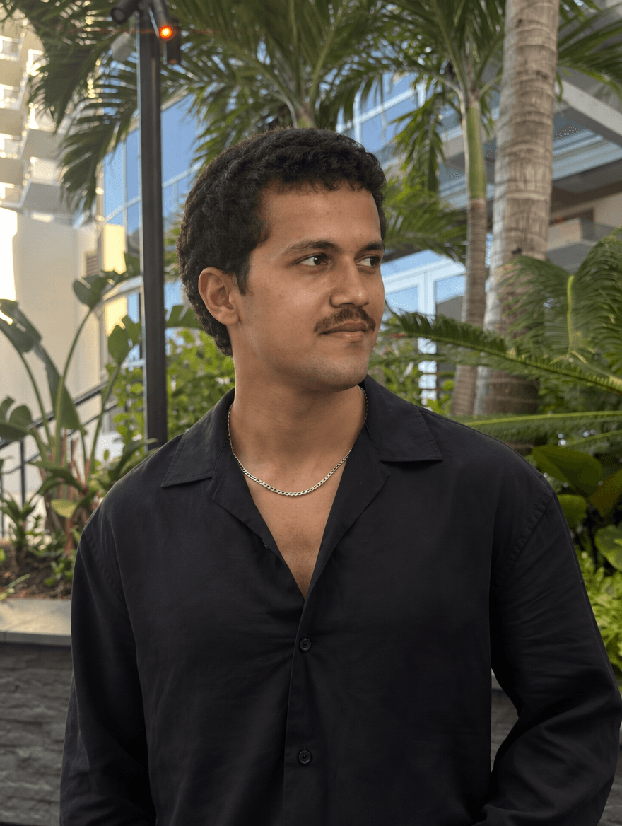 Man in a black linen shirt and silver chain among palm trees